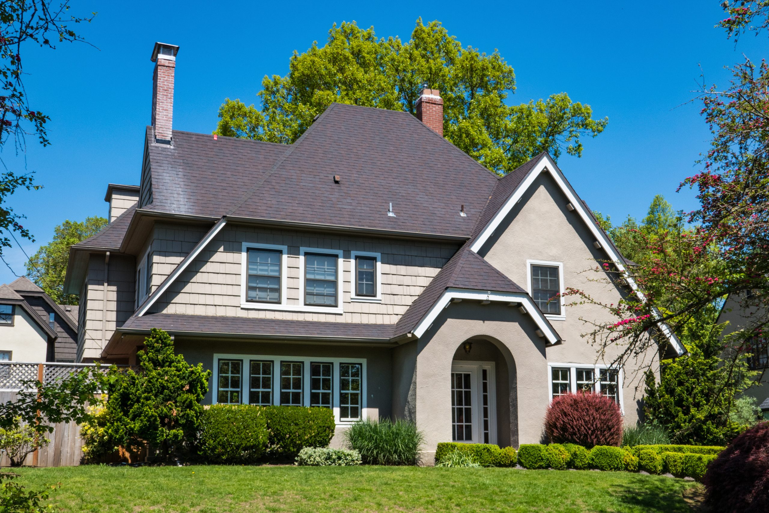 A two-story suburban house with gray siding and dark roof, arched entryway, multiple windows, and a well-maintained front yard with green grass, bushes, and trees under a clear blue sky.