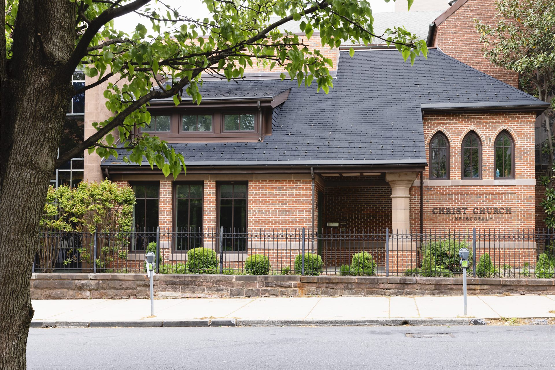 A brick building with arched windows and a sign reading "Christ Church Philadelphia" is surrounded by greenery and a black iron fence, seen from across a quiet street with parking meters and a tree in the foreground.