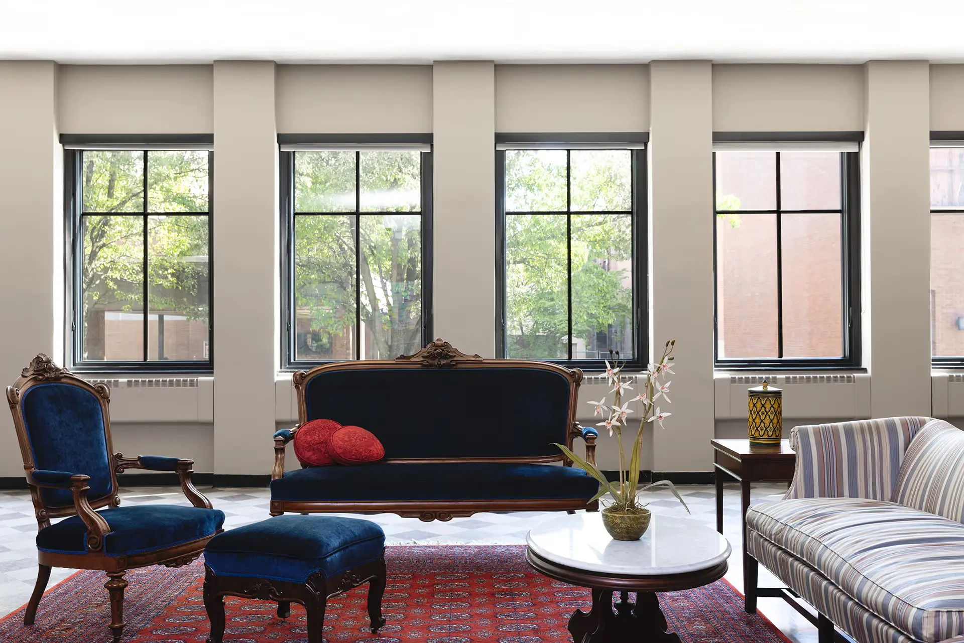 A bright sitting room with large windows, featuring a blue velvet chair and sofa, a striped couch, a red patterned rug, a round marble coffee table with a potted plant, and two red accent pillows.