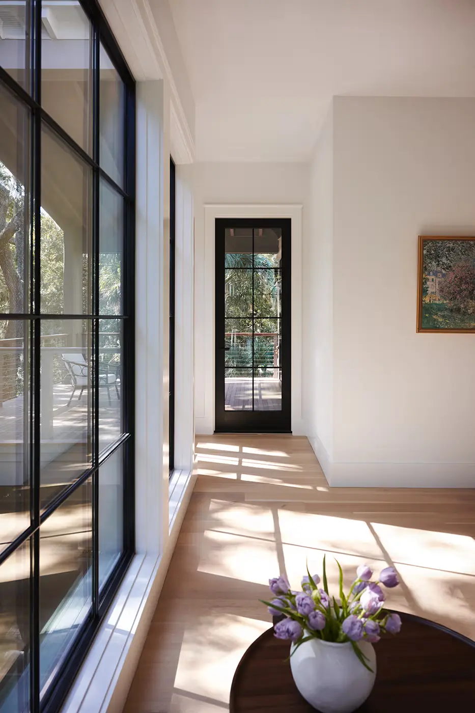 Bright hallway with large black-framed windows casting shadows on a light wood floor, a black-framed glass door at the end, a painting on the wall, and a round table with a vase of purple tulips in the foreground.