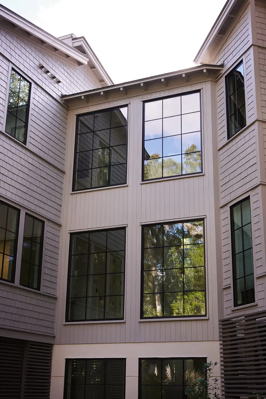 Three-story modern building with large grid-style windows reflecting trees and sky, surrounded by light gray shingles and white siding. The view is upward toward the roof and bright sky above.