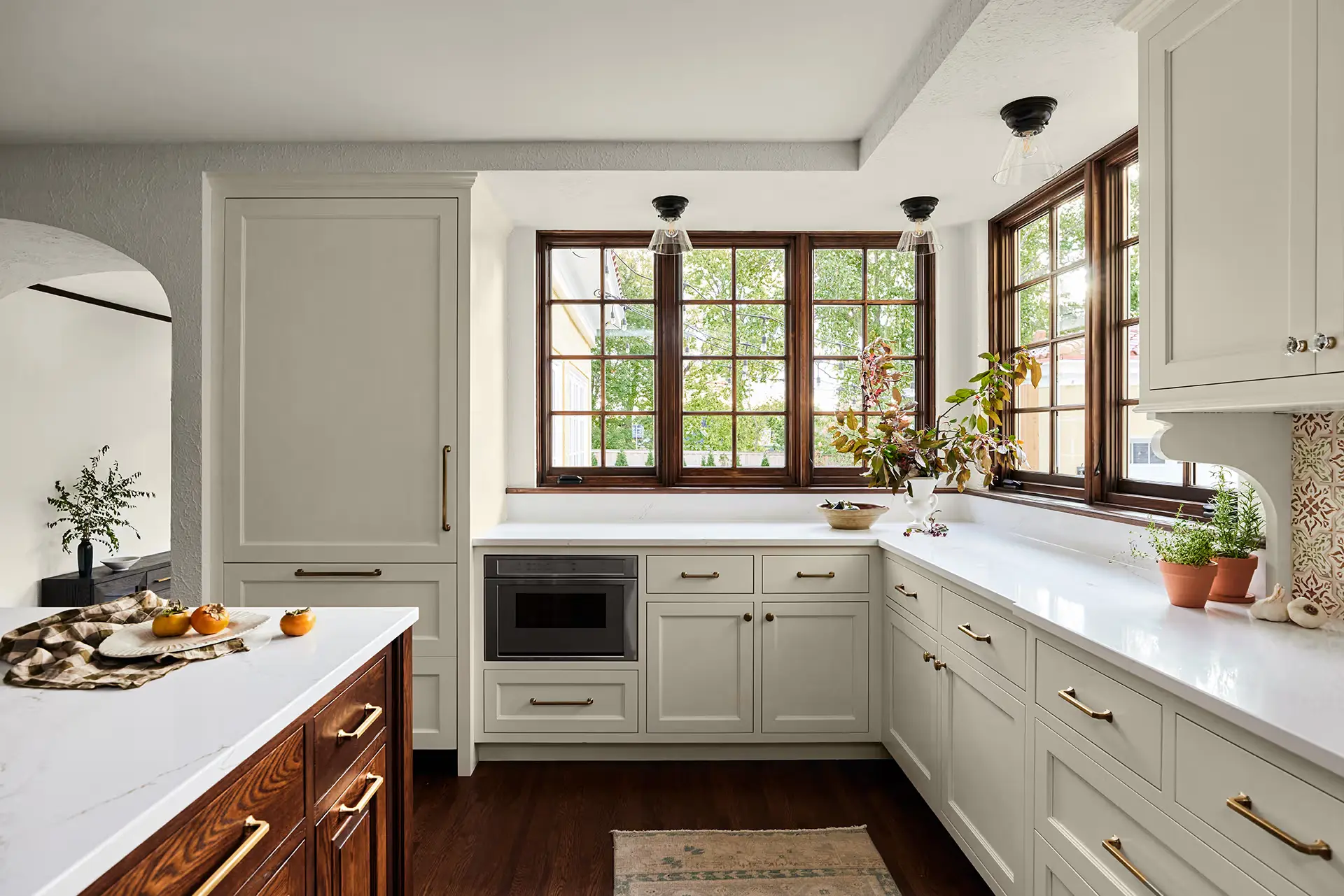 Bright kitchen with white cabinets, gold hardware, a built-in microwave, and large windows letting in natural light. Plants and decor sit on the counters, and there’s a wooden island and dark wood floor.