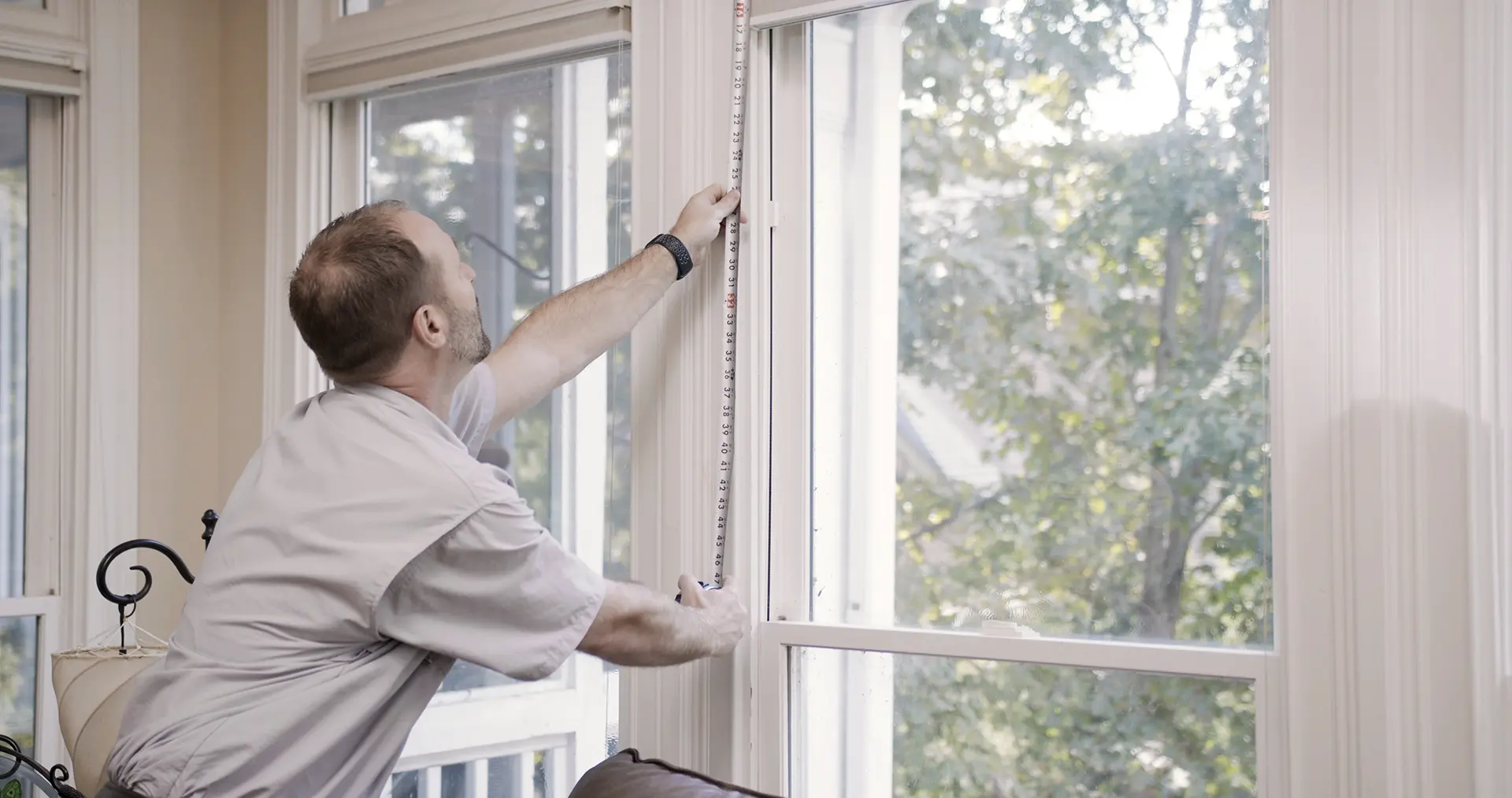MeasureOrder A person in a light gray shirt uses a tape measure to measure the height of a large window inside a bright room, with trees visible outside through the glass.