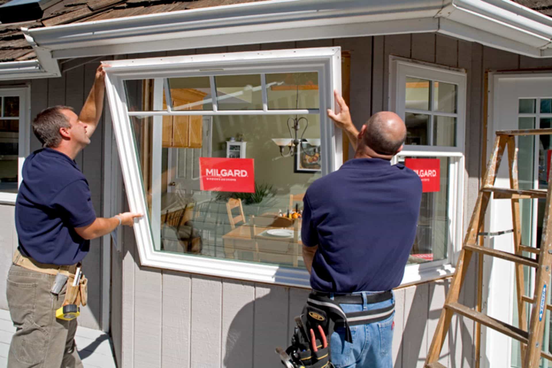 Two workers in blue shirts perform a historic window replacement, installing a large frame into the exterior wall of a house using tools and a wooden ladder. The interior dining room is visible through the opening.