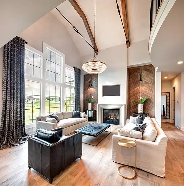 A bright, modern living room with high vaulted ceilings, exposed wooden beams, large windows, two white sofas, a black armchair, a central fireplace, and contemporary decor.