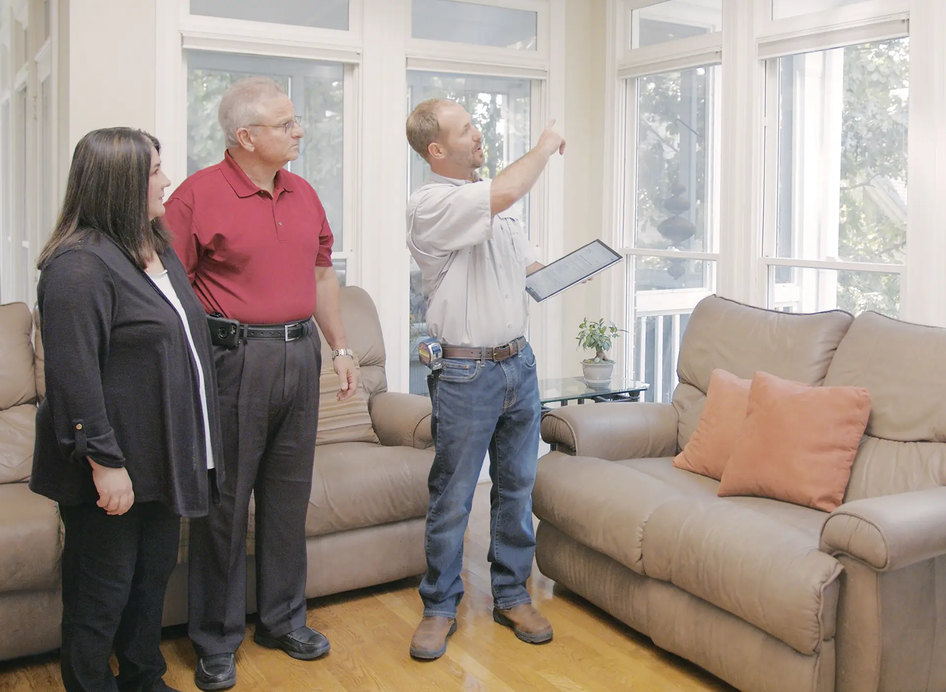 filtersquality100filltransparent A man in a white shirt holding a tablet points toward large windows while talking to an older couple in a living room with beige sofas and wooden floors.