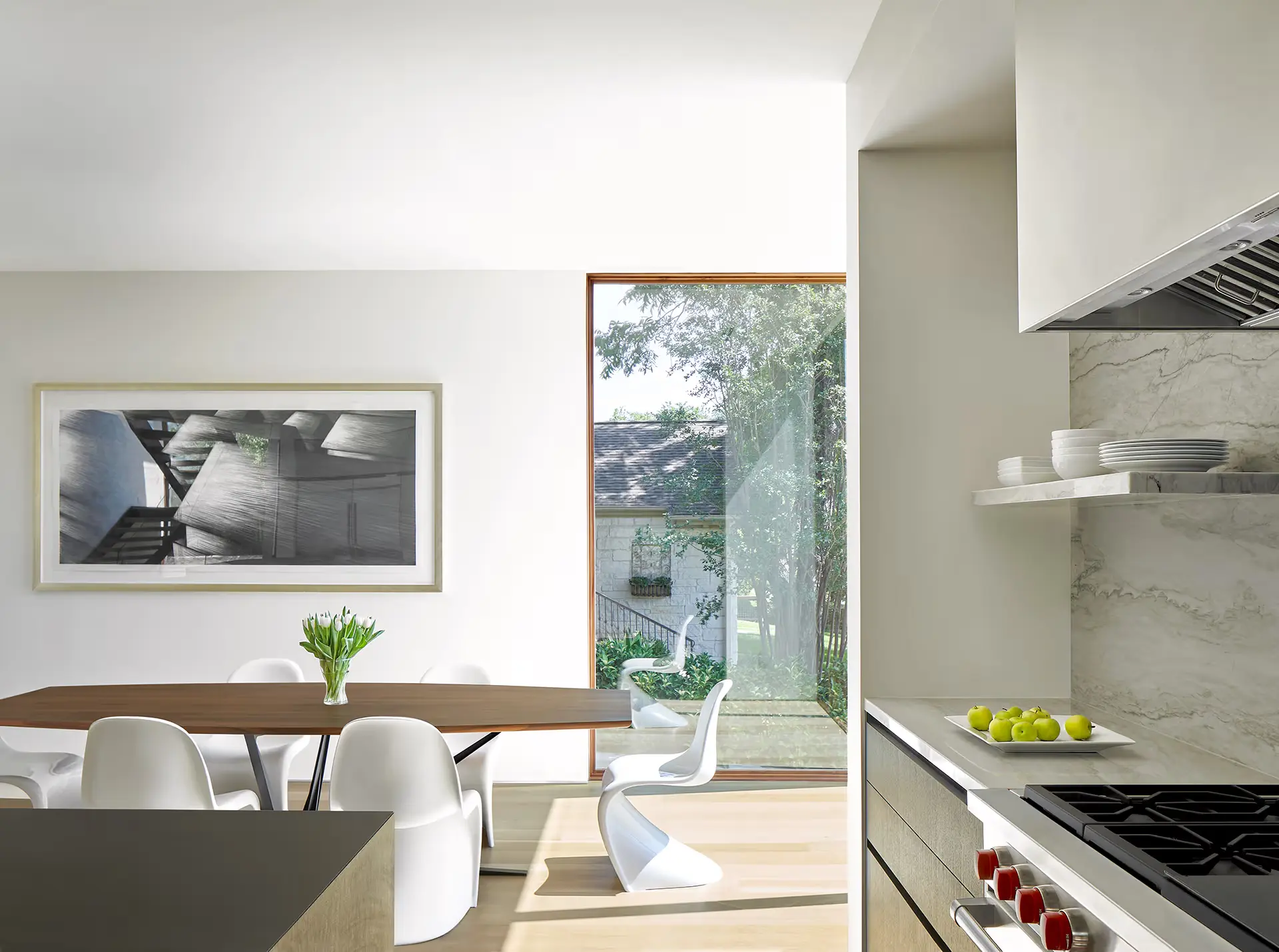 summer_heat Modern kitchen and dining area with white chairs around a wooden table, a vase of flowers, a large window showing greenery outside, open shelves with dishes, and a stove with red knobs in the foreground.