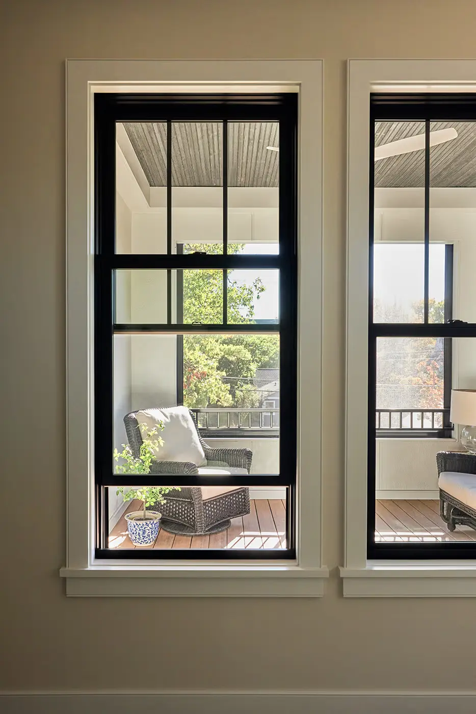 summer_heat_type View through two large windows showing a porch with wicker chairs, beige cushions, a small plant in a blue and white pot, and trees visible outside. Sunlight brightens the cozy seating area.