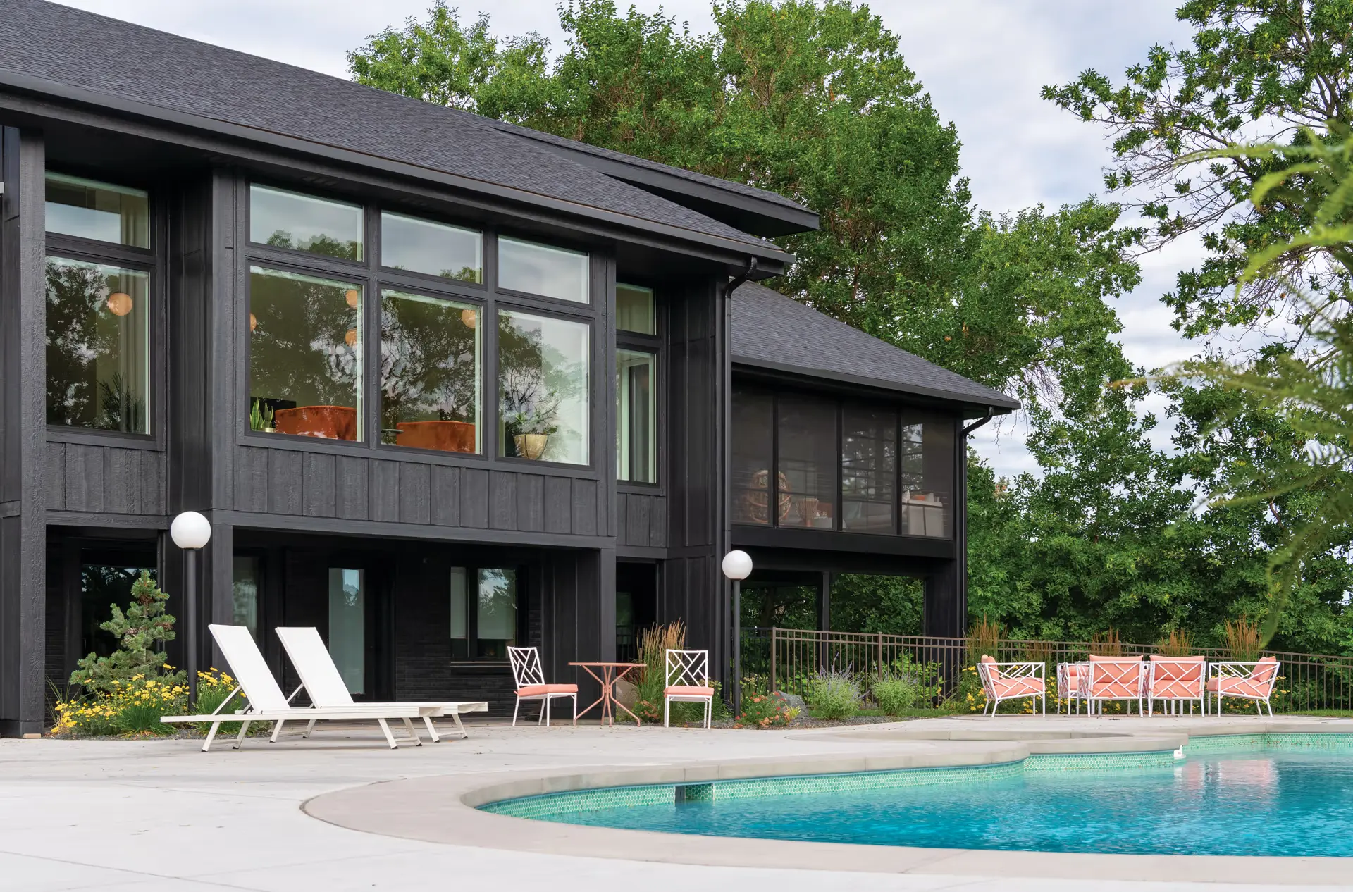Modern two-story black house with large windows featuring historic window replacement, poolside lounge chairs, a round swimming pool, and outdoor seating area, surrounded by trees and greenery on a cloudy day.