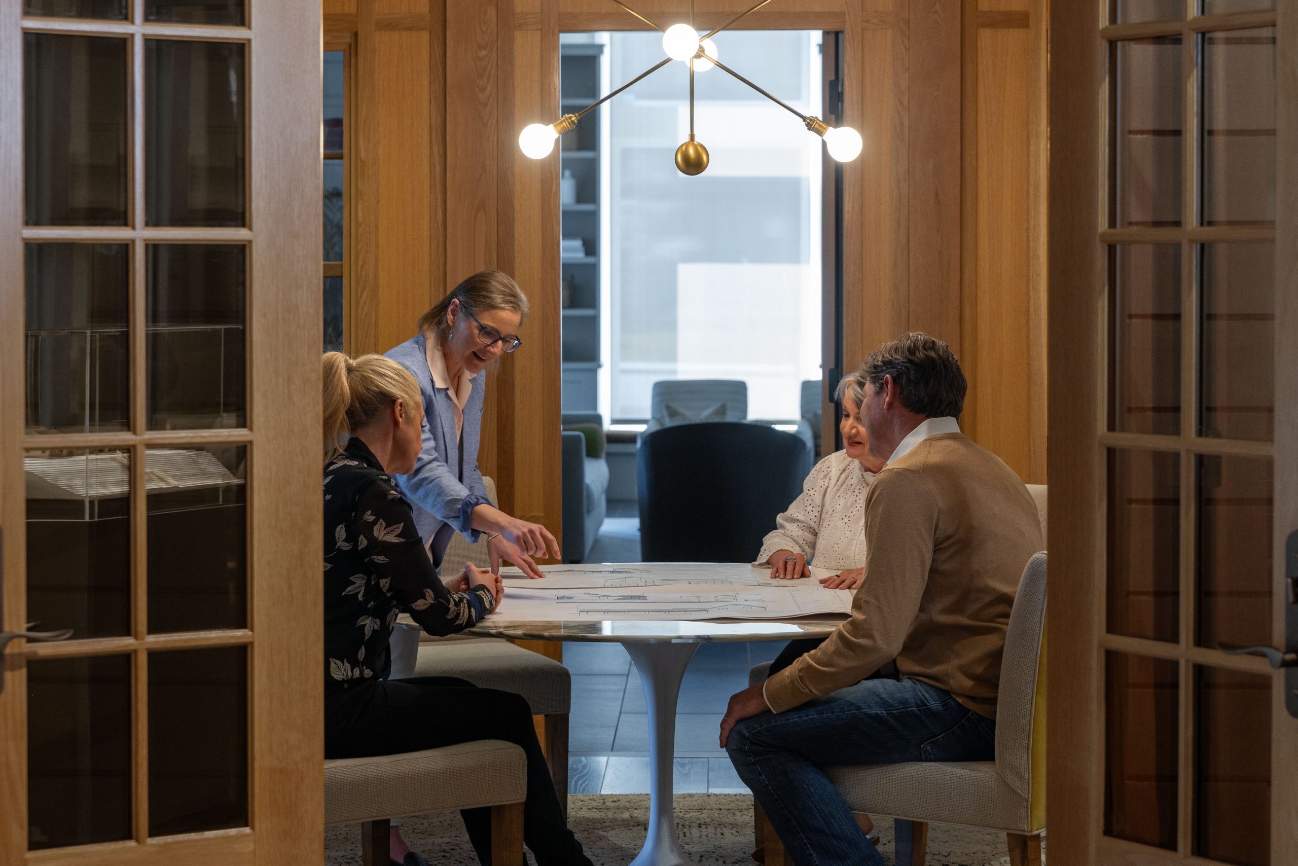 Four people sit around a round table in a modern office, reviewing documents together. One person stands, pointing at papers, while the others are seated, listening and looking at the materials. Warm lighting fills the room.