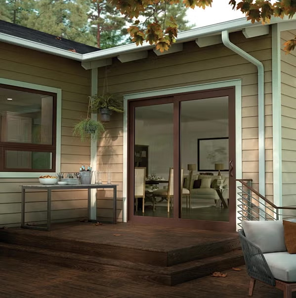A modern house with a wooden deck, glass sliding doors, and a window. A table with drinks and ice is on the deck, and a potted plant hangs above. Trees and outdoor furniture are visible.