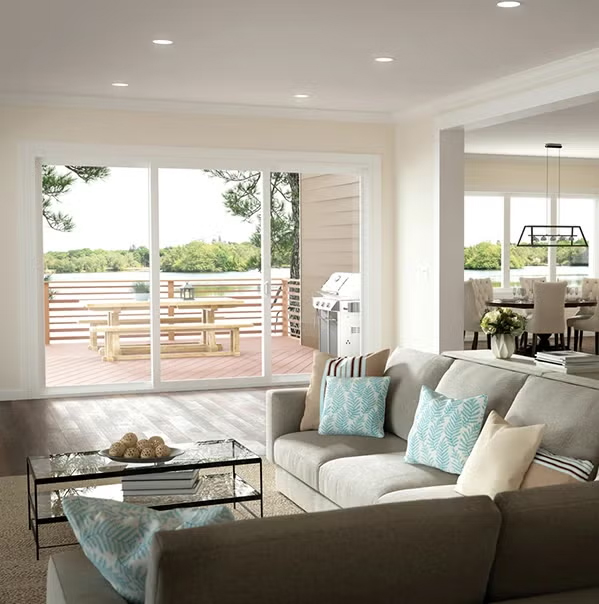 A bright living room with a gray sectional sofa and blue patterned pillows faces a glass coffee table. Large sliding glass doors open to a deck with a wooden picnic table and grill, overlooking green trees.