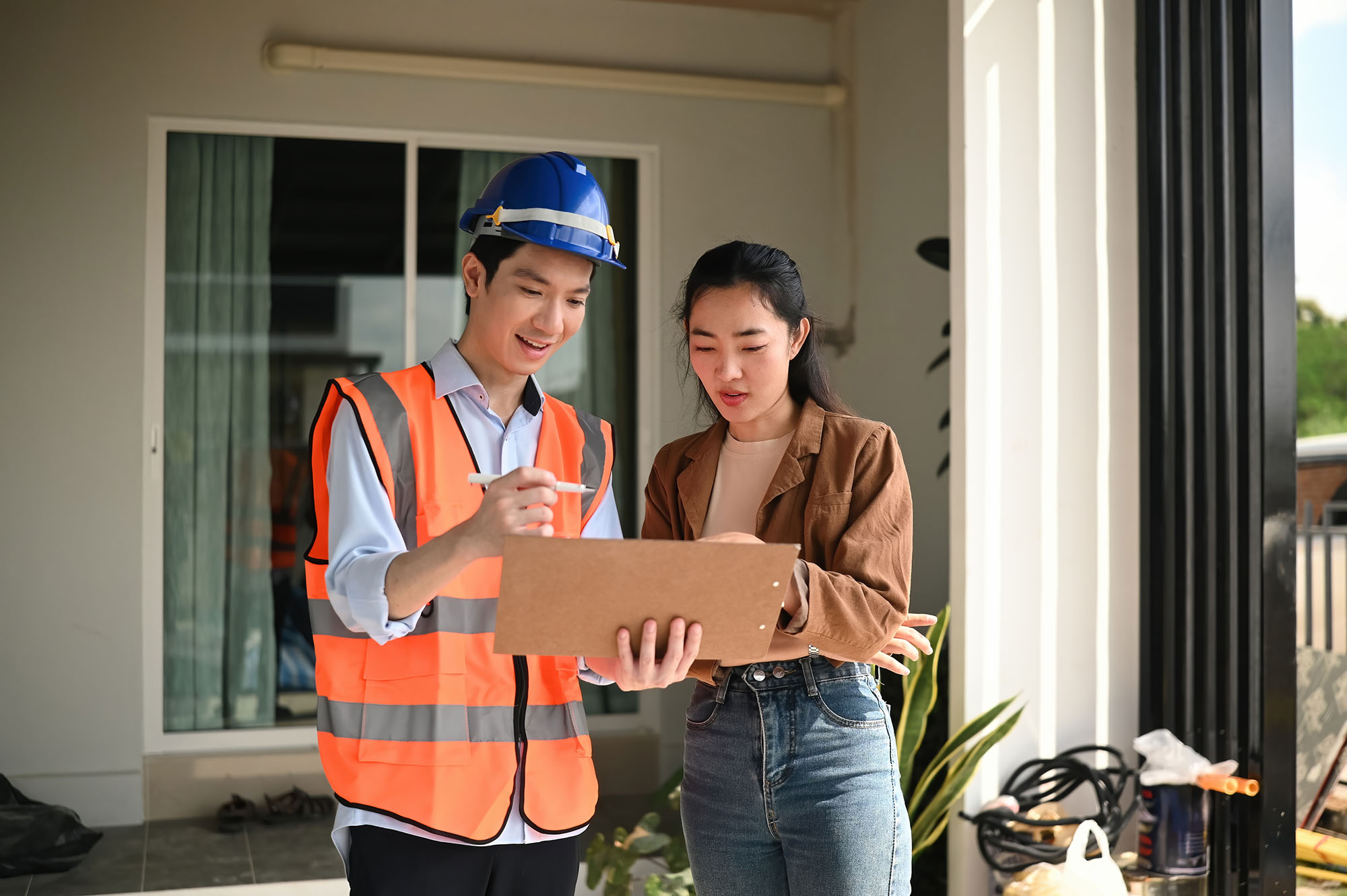 A construction worker in a safety vest and helmet shows a clipboard to a woman; they are standing outside a building, reviewing documents together.