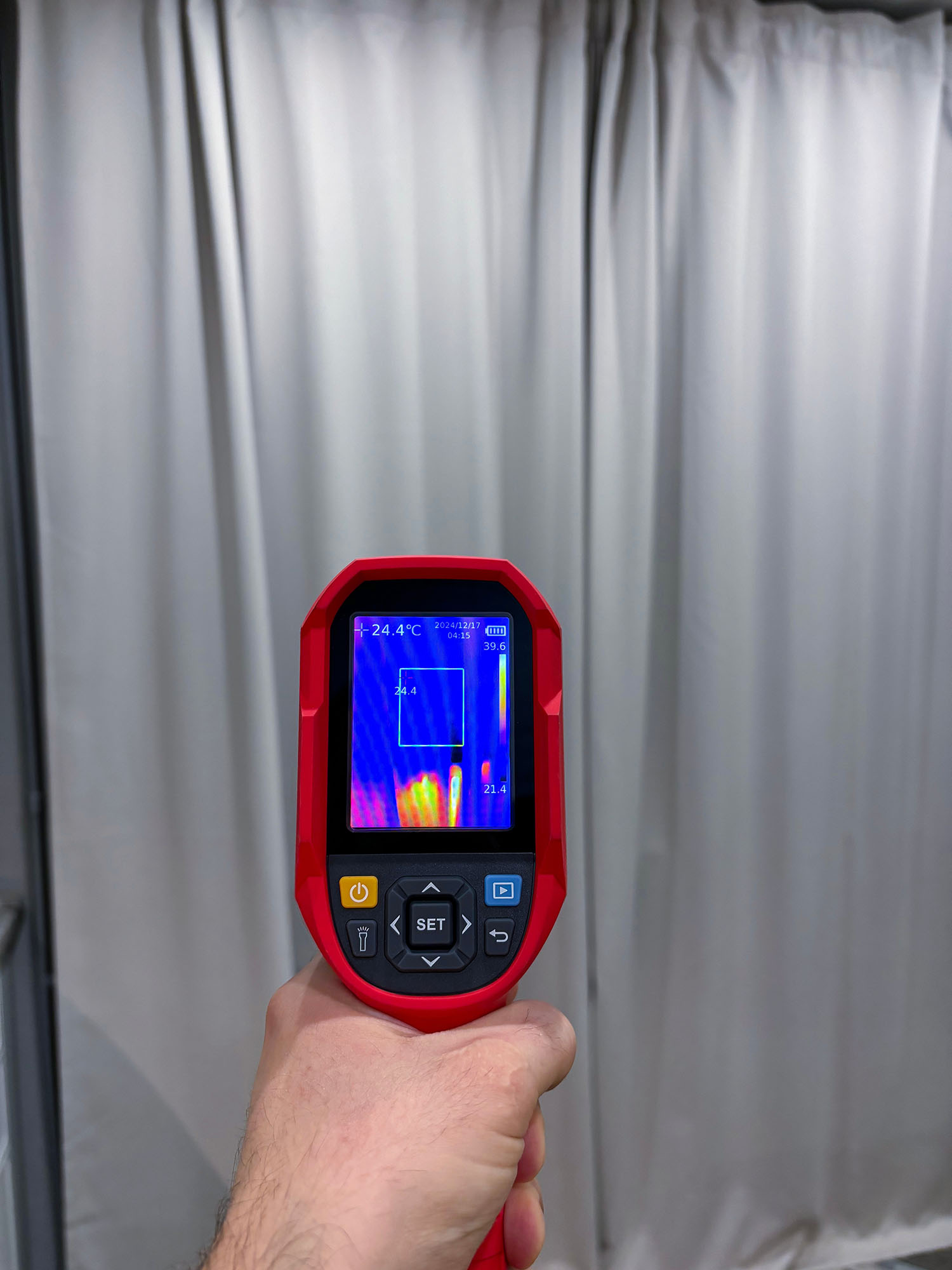 A hand holds a red thermal imaging camera aimed at white curtains. The camera screen shows a heat signature with colors ranging from blue to yellow, indicating temperature differences in the area in front of the curtains.