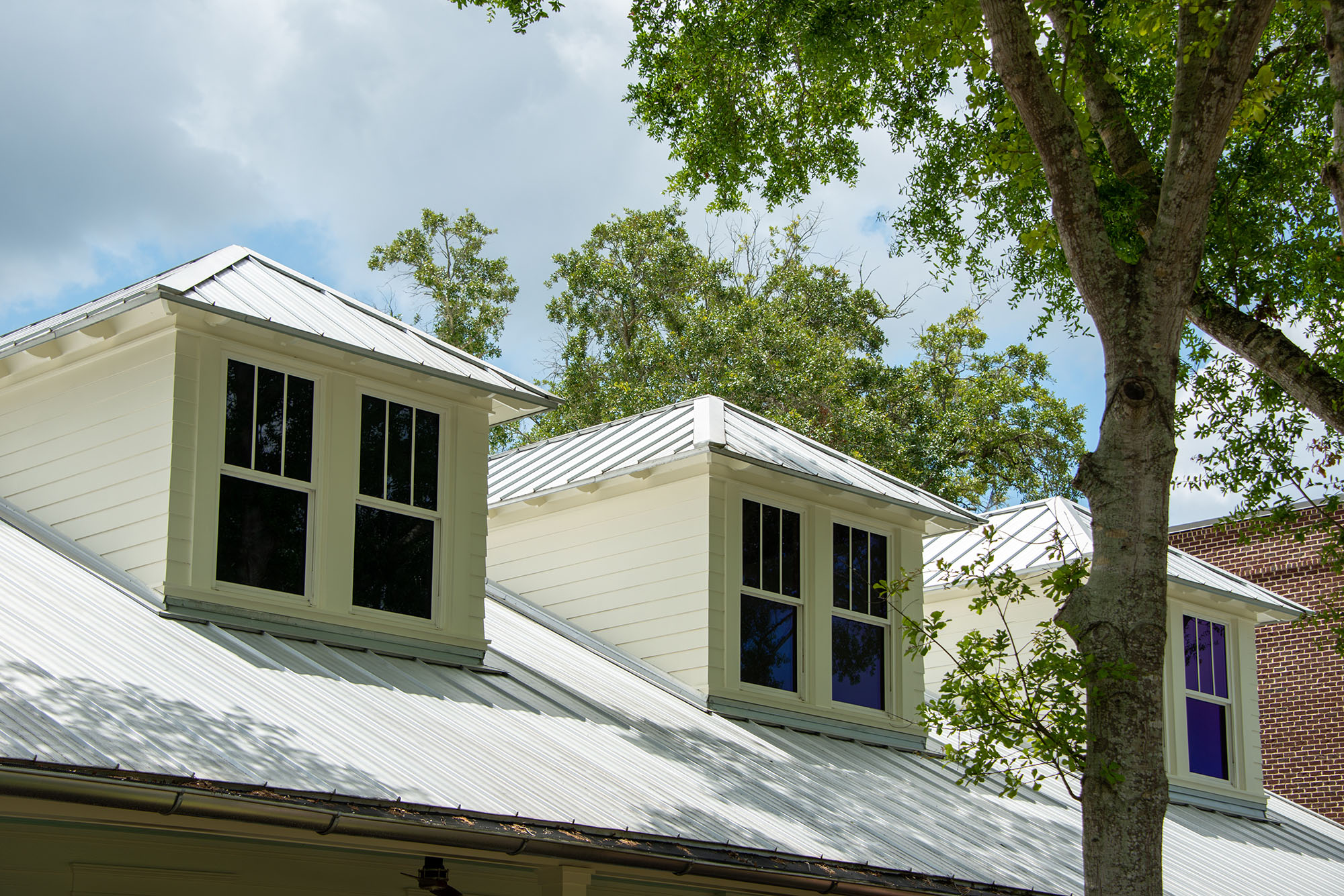 Three,Dormers,With,Multiple,Double,Hung,Windows,,A,Grey,Metal A white house with a metal roof and two dormer windows, surrounded by green trees under a partly cloudy sky.